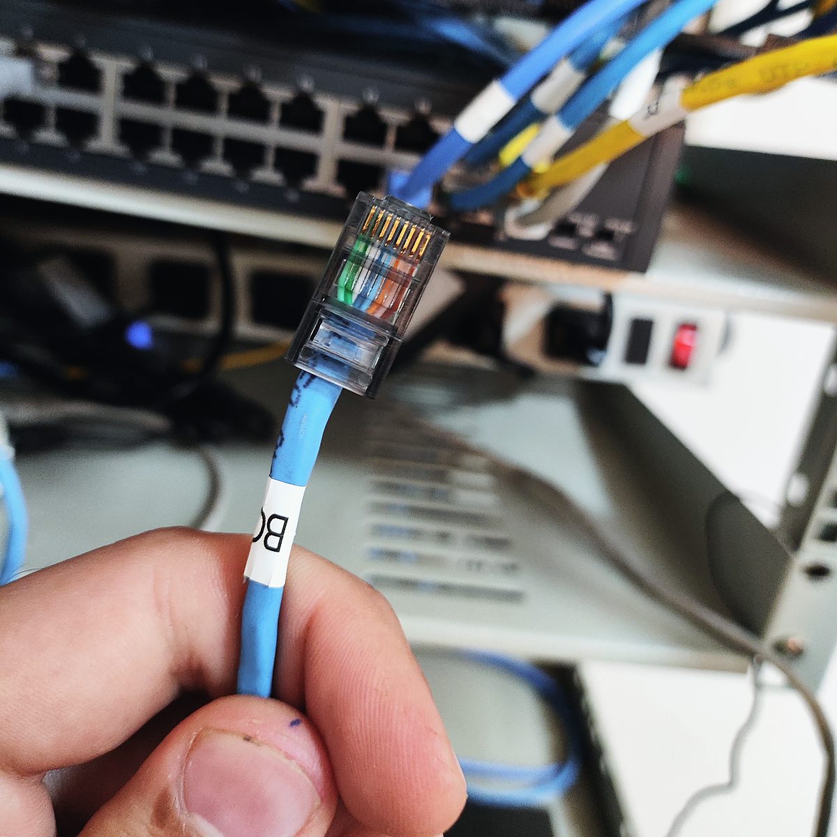 Mão segurando um cabo de rede azul com plugue RJ45 e etiqueta de identificação, diante de um patch panel