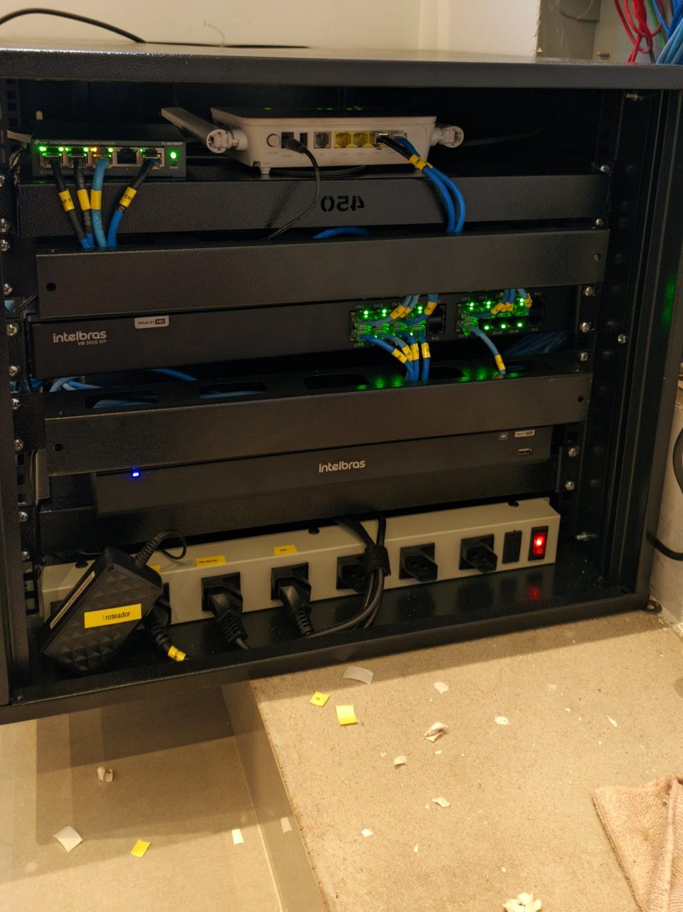 Rack de parede aberto com switch, roteador, equipamentos Intelbras de CFTV e cabos azuis identificados
