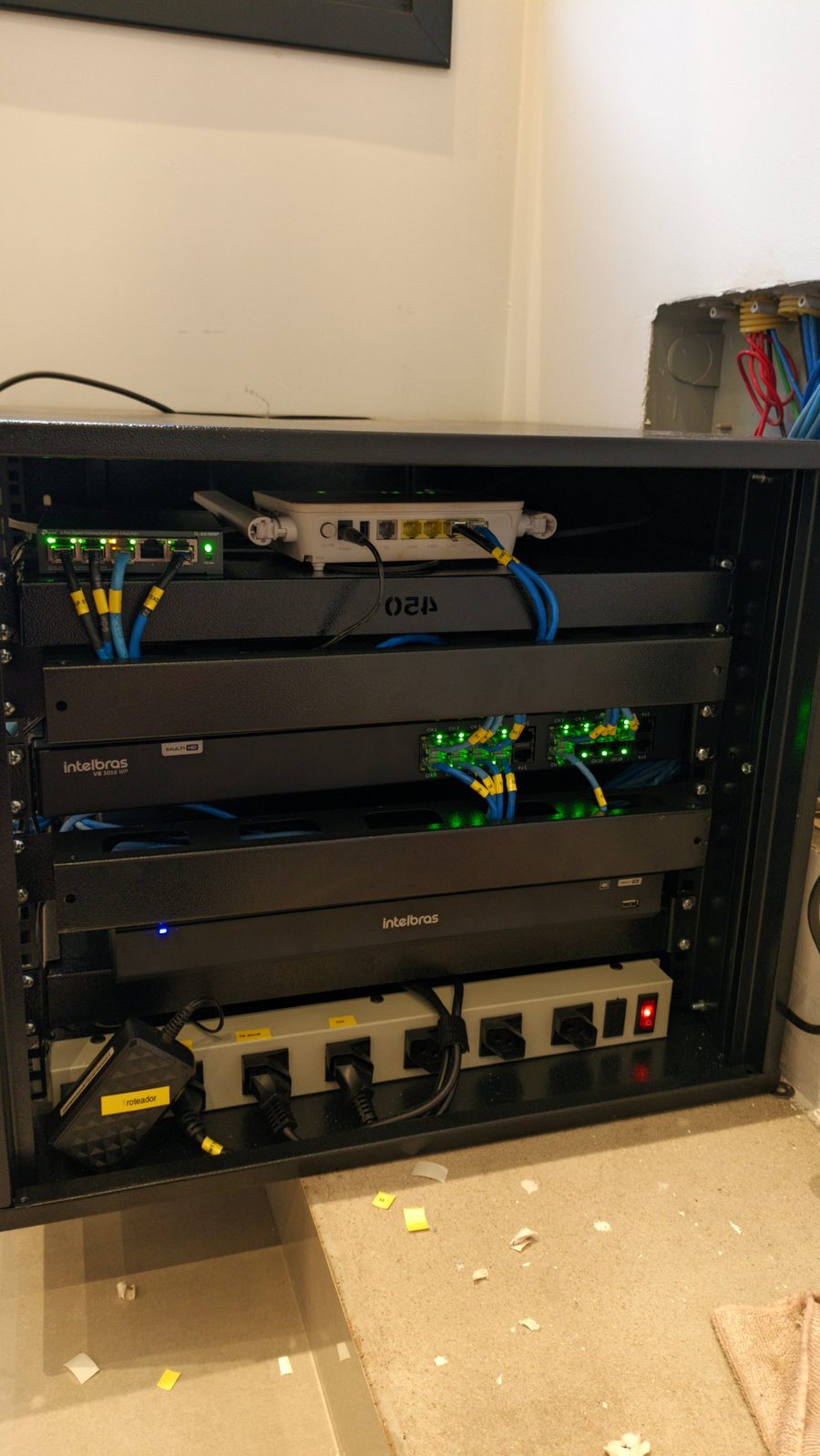 Rack de parede organizado com switch PoE, roteador, gravadores Intelbras, patch panels e cabos azuis identificados