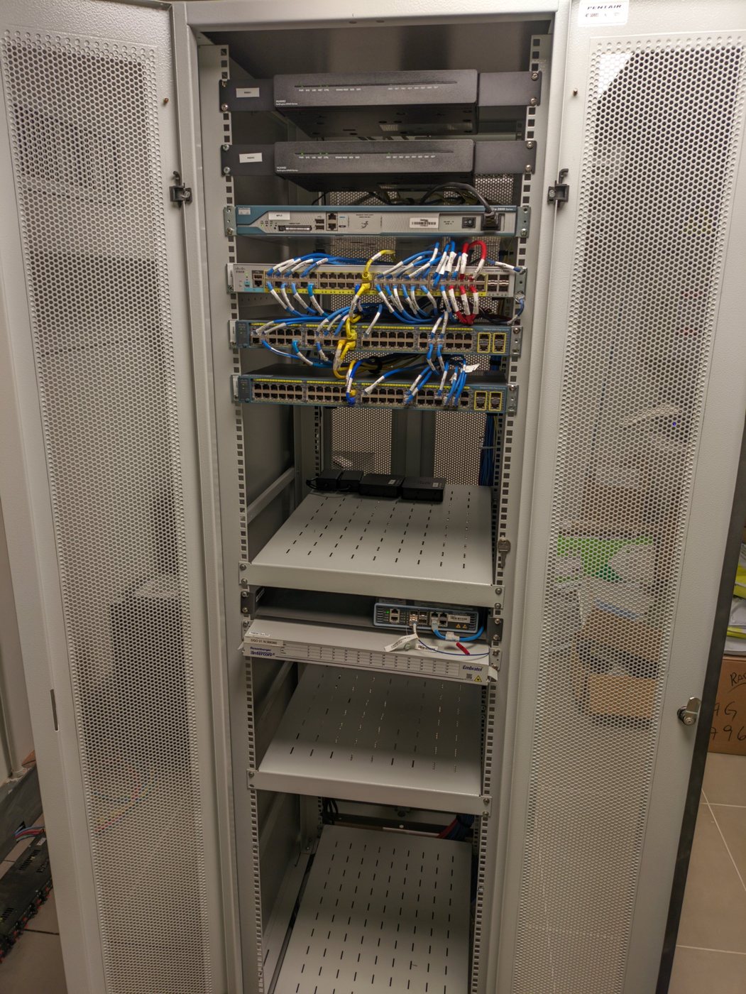 Rack de rede aberto, com roteadores, switches, cabos azuis identificados, patch panel de fibra e bandejas
