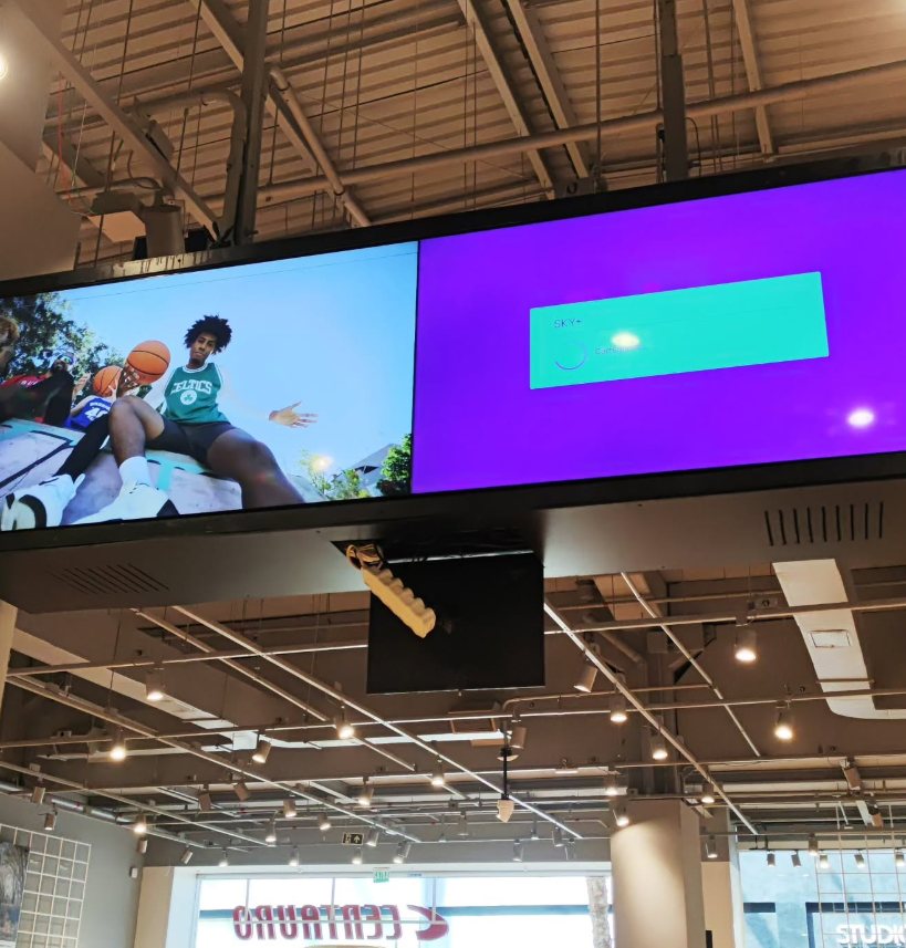 Painel de TV largo suspenso no teto de uma loja, exibindo conteúdo em duas telas